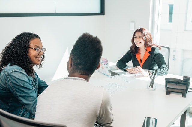 A woman of color wearing professional business attire sits behind a desk with a computer and other desk accessories in front of a couple of color having a conversation.