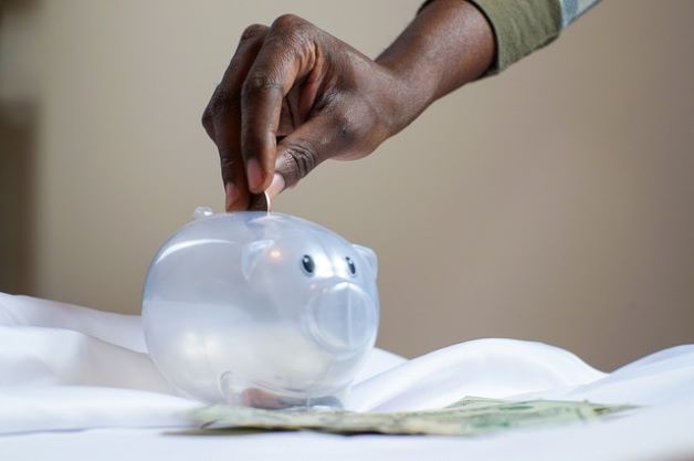 The hand of a person of color places a coin in a clear plastic piggy bank in the form of a pig, as the blurry shape of paper bills appear to the right side of the photo.