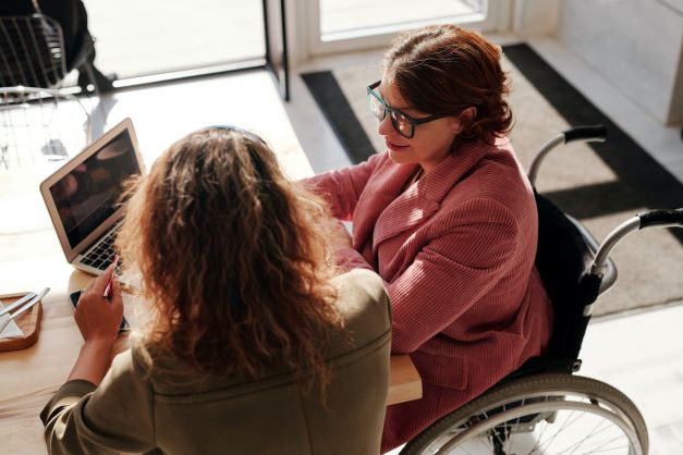 Woman who uses a wheelchair sits in front of a computer with another person sitting to her side.