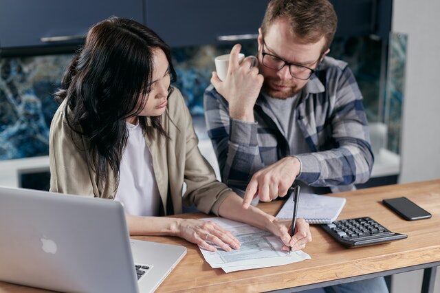 A man with a drinking mug in his hand, wears a plaid shirt and points to an unidentified document. A woman is sitting next to him wearing a beige shirt. A calculator, cell phone, and computer are placed in front of them while sitting at a desk as the woman completes the form  using a black pen.