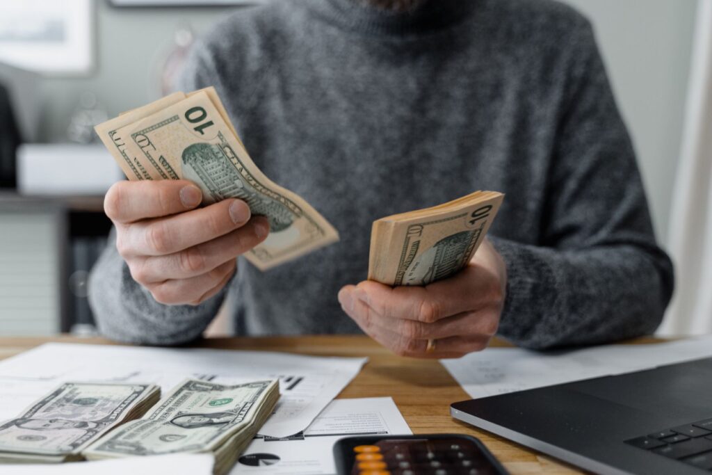 A person wearing a gray turtleneck sweater holds a pile of ten dollar bills in each hand, and two piles of bills of different denominations are placed in front of them, with a computer and calculator set off to the side, as though they are counting money and calculating bills.