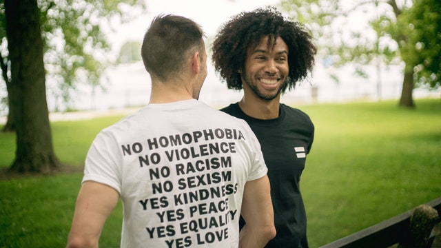 Two men, including a man of color facing forward, and a man with light colored skin facing away wears a t-shirt that reads: no homophobia, no violence, no racism, no sexism, yes kindness, yes peace, yes equality, yes love, appear to stand in a park among trees.